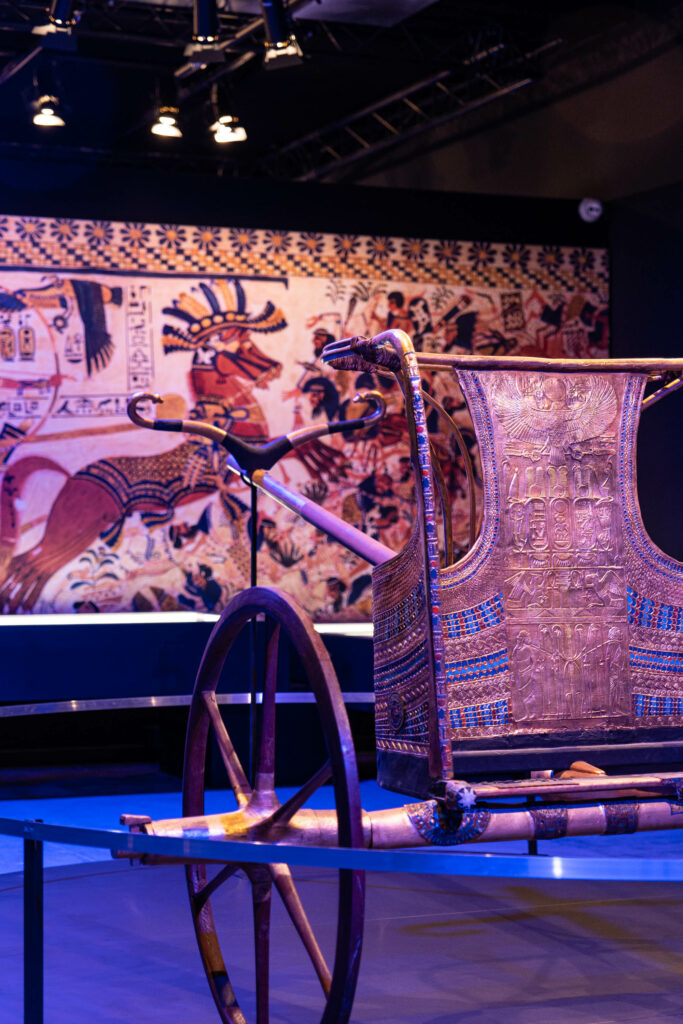 Une vue de l'exposition Toutankhamon à Metz : on y voit au premier plan une reconstitution d'un char égyptien d'époque et en fond, une toile dorée avec des dessins évoquant la civilisation égyptienne. Parfait pour offrir un cadeau !